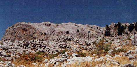 La cumbre de sierra Almola se levanta más allá del lapiaz del primer plano como la cabeza de un cachalote pétreo. A la derecha hiedras y encinas rupícolas en los acantilados.